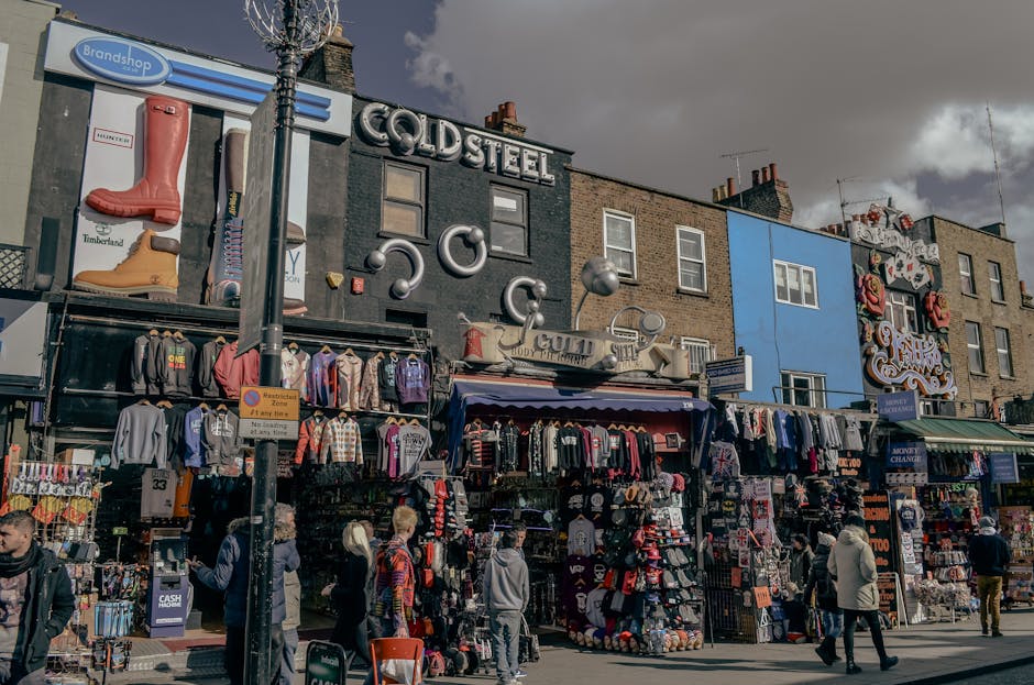 A busy outdoor scene at Camden Market in North London during daytime, with a prominent sign reading 'Camden Market' hanging above a crowd of people walking along the market street. The foreground feat